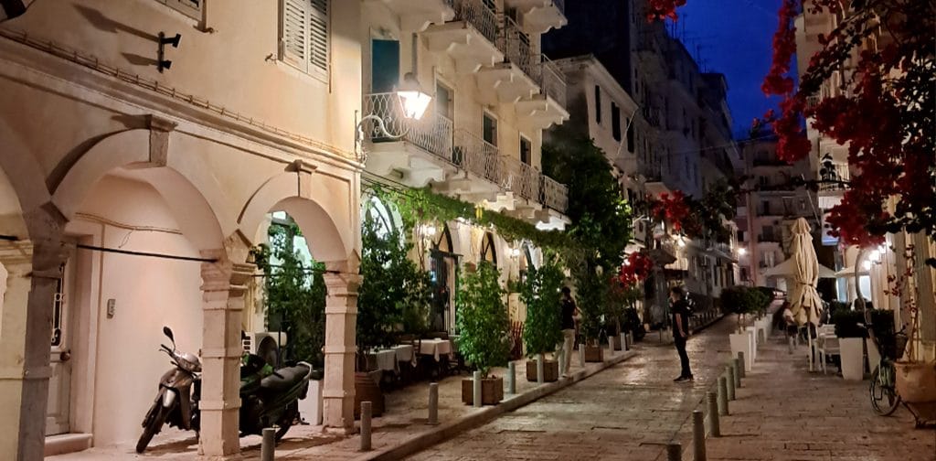 Narrow stone alley in Corfu Town's Old Town at night, with bougainvillea overhead and outdoor restaurant tables under Venetian-style lanterns