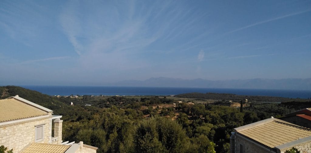 View from Northern Corfu over olive groves and tiled villa roofs to the Ionian Sea, with the mainland mountains in the distance